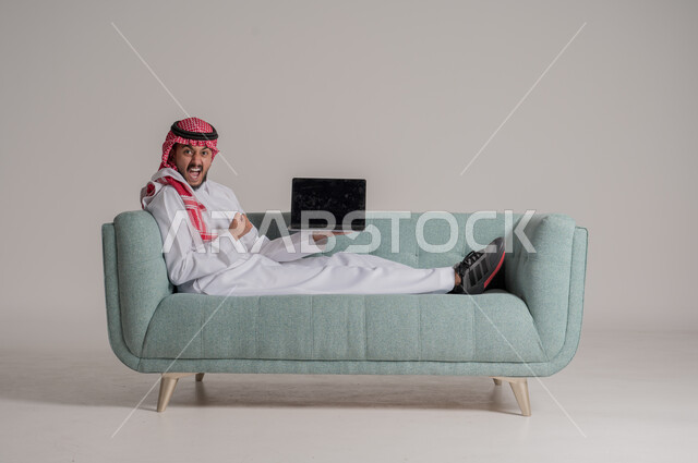 عرض شاشة إعلانية سوداء فارغة، استخدام الأجهزة والتقنيات الحديثة، رفع اليد للاعلى بإيماءات الانفعال والسرور بتطبيقات الجهاز اللوحي، بورتريه لرجل عربي خليجي سعودي يرتدي الزي التقليدي يمسك اللابتوب بتعابير السعادة، التسويق الرقمي الالكتروني، خلفية رمادية