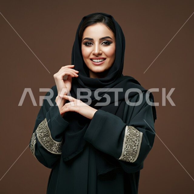 ايماءات السعادة والسرور، مفهوم النعومة والأنوثة، الشياكة والأناقة والاعتناء بالمظهر الخارجي، بورتريه مقرب لأمراة عربية خليجية سعودية محجبة مبتسمة ترتدي العباءة السوداء تنظر الى الكاميرا بتعابير الفرح، خلفية بني