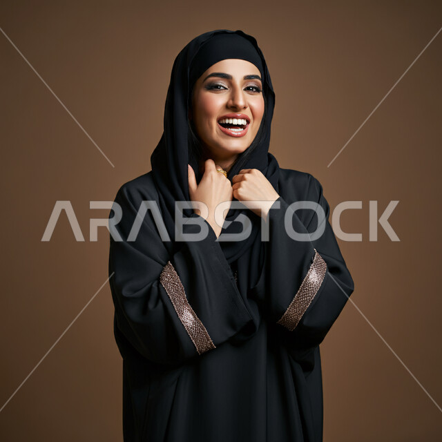 ايماءات السعادة والسرور، مفهوم النعومة والأنوثة، الشياكة والأناقة والاعتناء بالمظهر الخارجي، بورتريه مقرب لأمراة عربية خليجية سعودية محجبة مبتسمة ترتدي العباءة السوداء تنظر الى الكاميرا بتعابير الفرح، خلفية بني