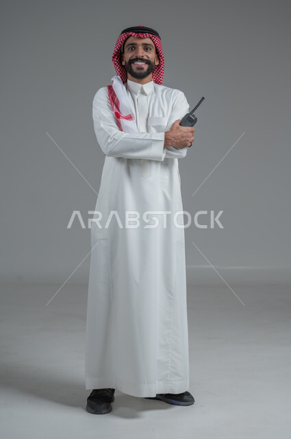 الإستجابة لحالات الطوارئ، المتابعة والإشراف على خطط العمل، بورتريه  لرجل عربي خليجي سعودي مبتسم يرتدي الثوب التقليدي والشماغ يقوم باستخدم جهاز الارسال والاستقبال اللاسلكي، مفهوم ادارة المشاريع عن بعد، صورة لكامل الجسم، خلفية رمادية