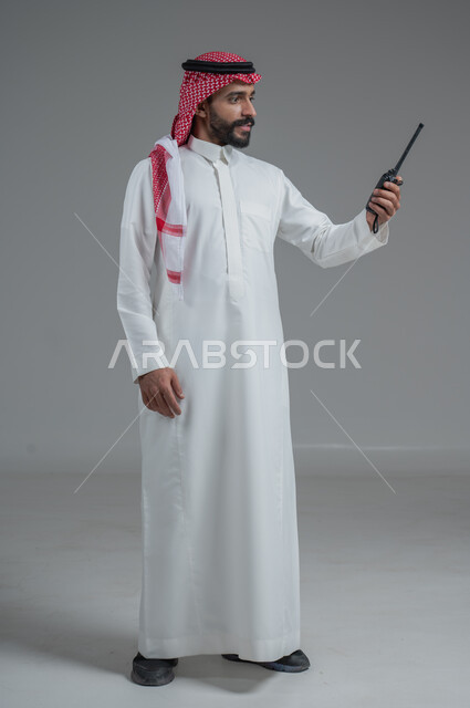 الإستجابة لحالات الطوارئ، المتابعة والإشراف على خطط العمل، بورتريه  لرجل عربي خليجي سعودي مبتسم يرتدي الثوب التقليدي والشماغ يقوم باستخدم جهاز الارسال والاستقبال اللاسلكي، مفهوم ادارة المشاريع عن بعد، صورة لكامل الجسم، خلفية رمادية