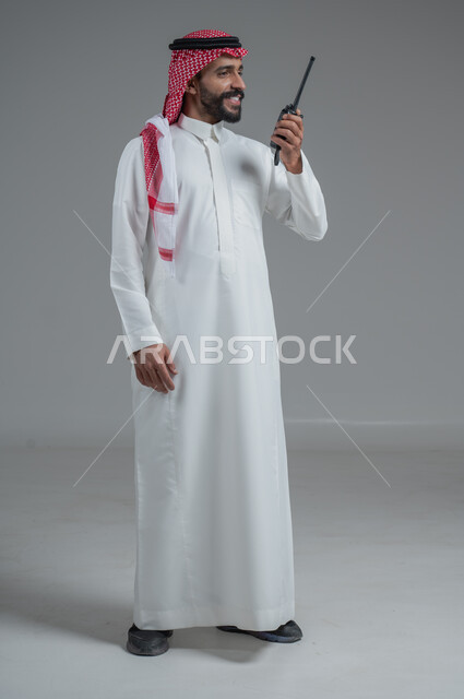 الإستجابة لحالات الطوارئ، المتابعة والإشراف على خطط العمل، بورتريه  لرجل عربي خليجي سعودي مبتسم يرتدي الثوب التقليدي والشماغ يقوم باستخدم جهاز الارسال والاستقبال اللاسلكي، مفهوم ادارة المشاريع عن بعد، صورة لكامل الجسم، خلفية رمادية