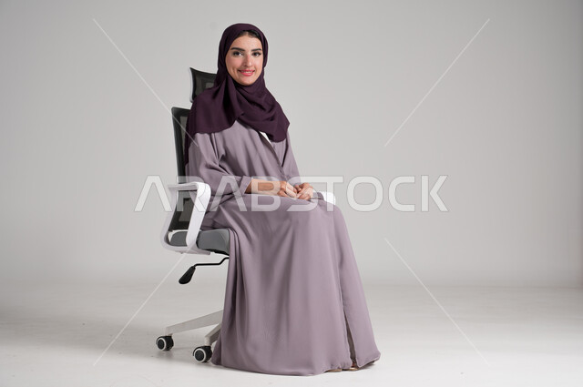 مفهوم الأناقة والأنوثة، بورتريه لامرأة عربية خليجية سعودية محجبة ترتدي العباءة التقليدية تجلس على كرسي دوار باتزان وهدوء وتنظر إلىالكاميرا بإيماءات السرور والبشاشة، الملابس النسائية العصرية للمحجبات، الاهتمام بالمظهر الخارجي، الجسم بالطول الكامل، خلفية رمادية