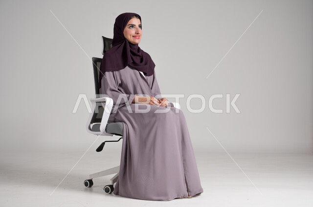 مفهوم الأناقة والأنوثة، بورتريه لامرأة عربية خليجية سعودية محجبة ترتدي العباءة التقليدية تجلس على كرسي دوار باتزان وهدوء وتنظر إلى مكان ما بإيماءات التفكير العميق، الملابس النسائية العصرية للمحجبات، الاهتمام بالمظهر الخارجي، الجسم بالطول الكامل، خلفية رمادية