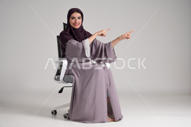 Pointing to a distant place, portrait of a veiled Saudi Arabian Gulf woman wearing a traditional abaya sitting on a swivel chair raising her hands and pointing in a certain direction, concept of elegance and femininity, modern women's clothing for veiled women, interest in external appearance, full-length body, gray background
