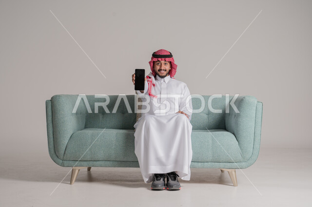 التسويق الرقمي الالكتروني، عرض شاشة سوداء فارغة، استخدام الأجهزة والتقنيات الحديثة، الإعجاب بتطبيقات جهاز الهاتف المحمول، بورتريه لرجل عربي خليجي سعودي يرتدي الثوب التقليدي والشماغ بجلس على كنبة مريحة يمسك الموبايل بتعابير السعادة، خلفية رمادية