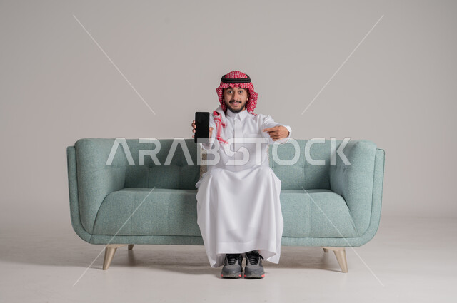 Black screen throne empty on mobile, looking at the camera and pointing to the mobile phone with expressions and gestures of admiration, portrait of a smiling Saudi Arabian Gulf man wearing a traditional thobe and a shemagh sitting on a sofa and using modern advanced technologies, gray background