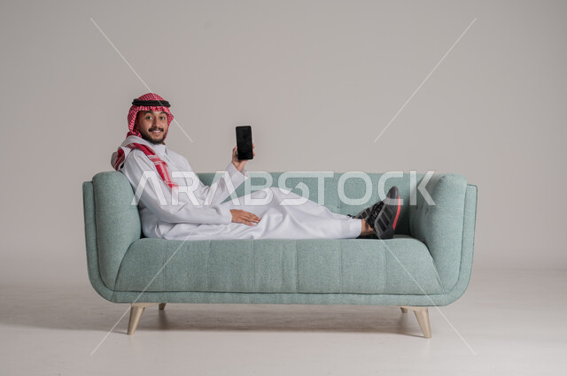 الإعجاب بتطبيقات جهاز الهاتف المحمول، بورتريه لرجل عربي خليجي سعودي يرتدي الثوب التقليدي والشماغ بستلقي على كنبة مريحة يمسك الموبايل بتعابير السعادة، التسويق الرقمي الالكتروني، عرض شاشة سوداء فارغة، استخدام الأجهزة والتقنيات الحديثة، خلفية رمادية