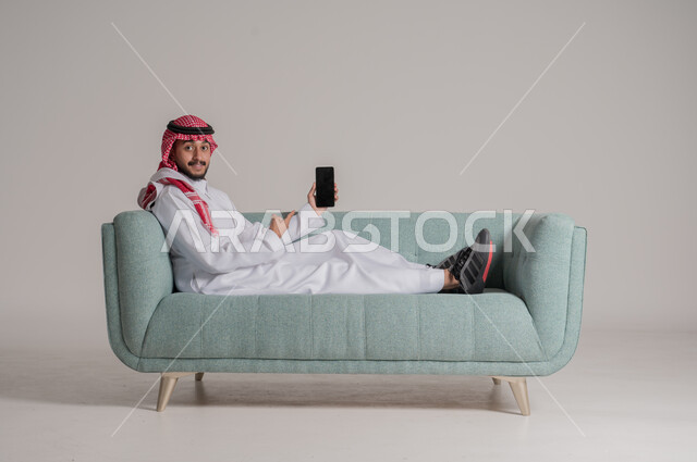 النظر الى الكاميرا والاشارة الى الهاتف المحمول المحمول بتعابير وايماءات الاعجاب، بورتريه لرجل عربي خليجي سعودي مبتسم يرتدي الثوب التقليدي والشماغ يستقلي على كنبة ويقوم باستخدام التقنيات الحديثه المتطورة، عرش شاشة سوداء فارغة على الجوال، خلفية رمادية