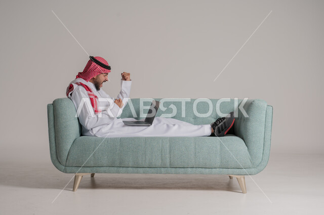 استخدام الأجهزة والتقنيات الحديثة، رفع اليدين للاعلى بإيماءات الانفعال والسرور بتطبيقات الجهاز اللوحي، بورتريه لرجل عربي خليجي سعودي يرتدي الزي التقليدي يمسك اللابتوب بتعابير السعادة، التسويق الرقمي الالكتروني، عرض شاشة سوداء فارغة، خلفية رمادية