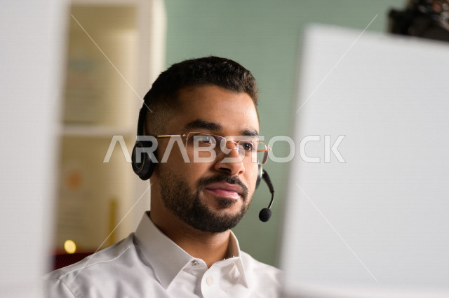 موظف عربي خليجي سعودي يعمل في خدمة العملاء ويرتدي سماعة الاتصال، استعلامات واستفسارات، الاجابه على اسئلة العملاء، اجراء اتصال فيديو من خلال الحاسوب، نظارة طبية، متابعة وتطوير العمل