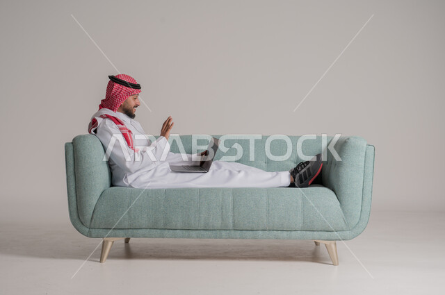 الدردشة مع الأهل والأصدقاء عبر الانترنت، استخدام الأجهزة الحديثة والمتطورة، إجراء مكالمة فيديو عبر وسائل التواصل الاجتماعي، بورتريه لرجل عربي خليجي سعودي يرتدي الثوب التقليدي والشماغ يستلقي على الكنبة ويحمل جهاز الحاسوب المحمول بايماءات السعادة، خلفية رمادية
