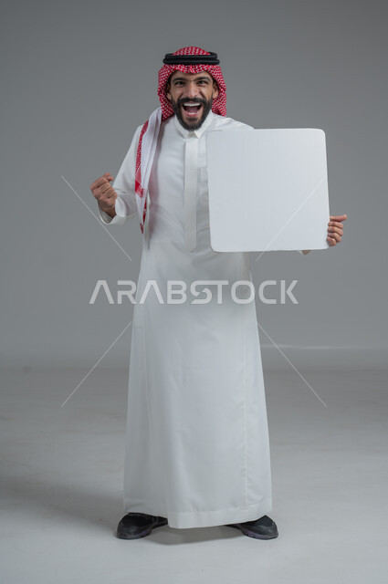 لوح إعلاني فارغ، موك اب أبيض، بورتريه لرجل عربي خليجي سعودي يرتدي الثوب التقليدي والشماغ يحمل بيده لوحه بيضاء، الوقوف باستقامه بايماءات الثقة بالنفس، صورة الجسم بالطول الكامل، خلفية رماديه