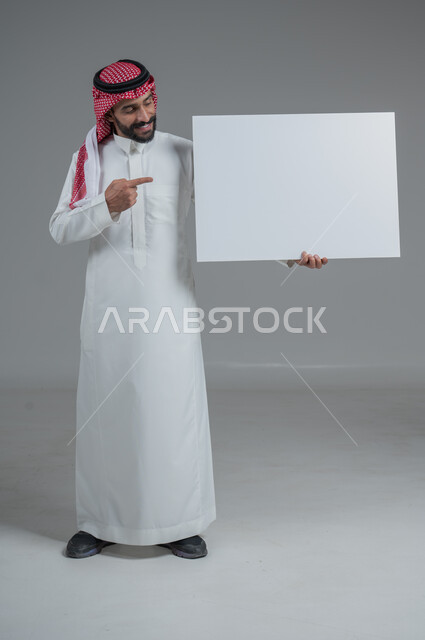 لوح إعلاني فارغ، موك اب أبيض، بورتريه لرجل عربي خليجي سعودي يرتدي الثوب التقليدي والشماغ يحمل بيده لوحه بيضاء، الوقوف باستقامه بايماءات الثقة بالنفس، صورة الجسم بالطول الكامل، خلفية رماديه