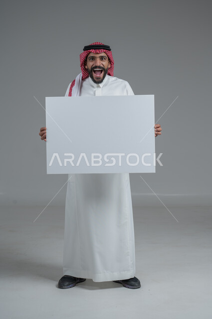 لوح إعلاني فارغ، موك اب أبيض، بورتريه لرجل عربي خليجي سعودي يرتدي الثوب التقليدي والشماغ يحمل بيده لوحه بيضاء، الوقوف باستقامه بايماءات الثقة بالنفس، صورة الجسم بالطول الكامل، خلفية رماديه