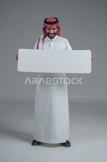 لوح إعلاني فارغ، موك اب أبيض، بورتريه لرجل عربي خليجي سعودي يرتدي الثوب التقليدي والشماغ يحمل بيده لوحه بيضاء، الوقوف باستقامه بايماءات الثقة بالنفس، صورة الجسم بالطول الكامل، خلفية رماديه