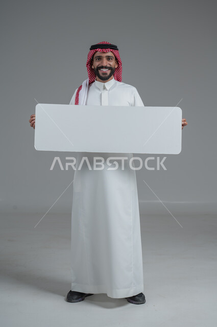 لوح إعلاني فارغ، موك اب أبيض، بورتريه لرجل عربي خليجي سعودي يرتدي الثوب التقليدي والشماغ يحمل بيده لوحه بيضاء، الوقوف باستقامه بايماءات الثقة بالنفس، صورة الجسم بالطول الكامل، خلفية رماديه