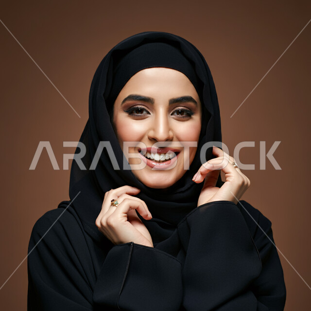 تعابير الفرح والسرور، الشياكة والأناقة والاعتناء بالمظهر الخارجي، بورتريه مقرب لأمراة عربية خليجية سعودية محجبة مبتسمة ترتدي العباءة السوداء تنظر الى الكاميرا بإيماءات السعادة والثقة بالنفس، مفهوم النعومة والأنوثة، خلفية بني
