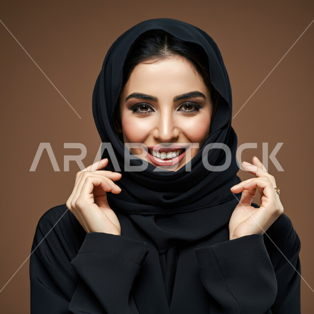 تعابير الفرح والسرور، الشياكة والأناقة والاعتناء بالمظهر الخارجي، بورتريه مقرب لأمراة عربية خليجية سعودية محجبة مبتسمة ترتدي العباءة السوداء تنظر الى الكاميرا بإيماءات السعادة والثقة بالنفس، مفهوم النعومة والأنوثة، خلفية بني