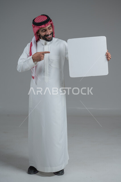 موك اب أبيض، لوح إعلاني فارغ،  بورتريه لرجل عربي خليجي سعودي يرتدي الثوب التقليدي والشماغ يحمل بيده لوحه بيضاء، الوقوف باستقامه بايماءات الثقة بالنفس، صورة الجسم بالطول الكامل، خلفية رماديه