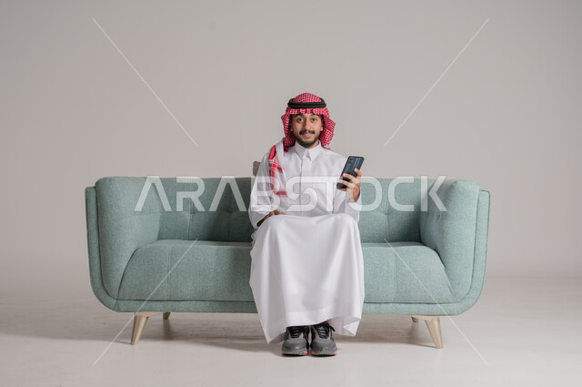 عقد إجتماع عمل عبر الإنترنت، استخدام الهاتف المحمول لإنجاز المهام، بورتريه لرجل عربي سعودي خليجي يرتدي الثوب التقليدي والشماغ يجلس على كنبة مريحة ويحمل جهاز الموبايل، النظر الى الكاميرا بايماءات السرور، تصفح برامج التواصل الاجتماعي, خلفية رمادية