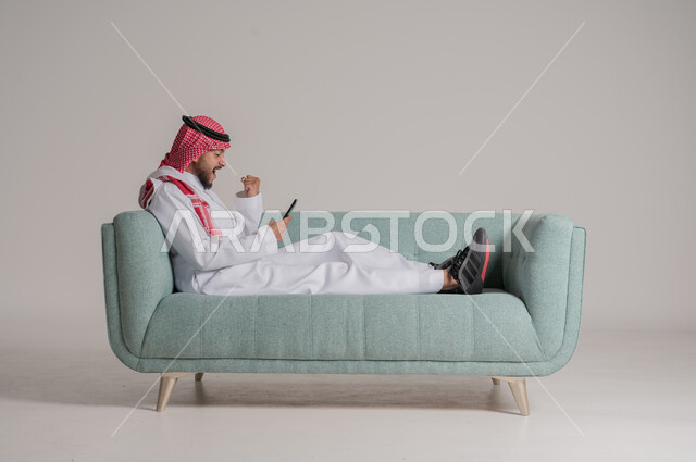 Raising hand up with gestures of excitement and pleasure with mobile applications, portrait of a Saudi Arabian Gulf man wearing traditional dress holding a mobile phone with expressions of happiness, electronic digital marketing, displaying a blank black screen, using modern devices and technologies, grey background