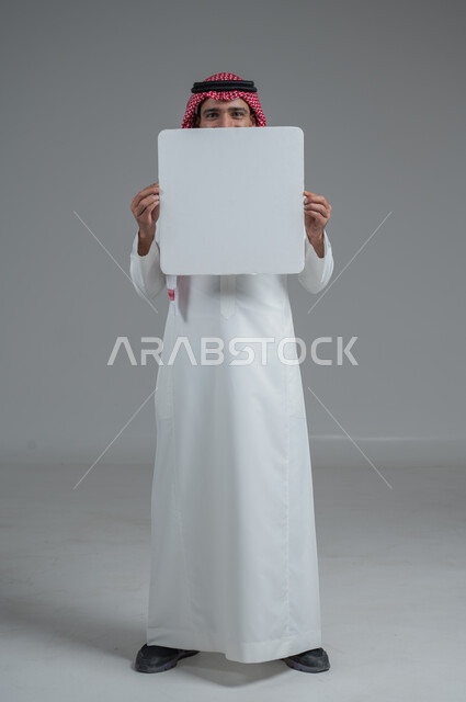 موك اب أبيض، لوح إعلاني فارغ،  بورتريه لرجل عربي خليجي سعودي يرتدي الثوب التقليدي والشماغ يحمل بيده لوحه بيضاء، الوقوف باستقامه بايماءات الثقة بالنفس، صورة الجسم بالطول الكامل، خلفية رماديه
