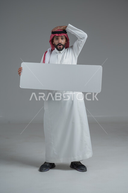موك اب أبيض، لوح إعلاني فارغ،  بورتريه لرجل عربي خليجي سعودي يرتدي الثوب التقليدي والشماغ يحمل بيده لوحه بيضاء بإيماءات الدهشة والصدمة، صورة الجسم بالطول الكامل، خلفية رماديه