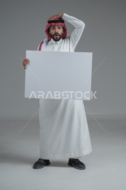 موك اب أبيض، لوح إعلاني فارغ،  بورتريه لرجل عربي خليجي سعودي يرتدي الثوب التقليدي والشماغ يحمل بيده لوحه بيضاء بإيماءات الدهشة والصدمة، صورة الجسم بالطول الكامل، خلفية رماديه