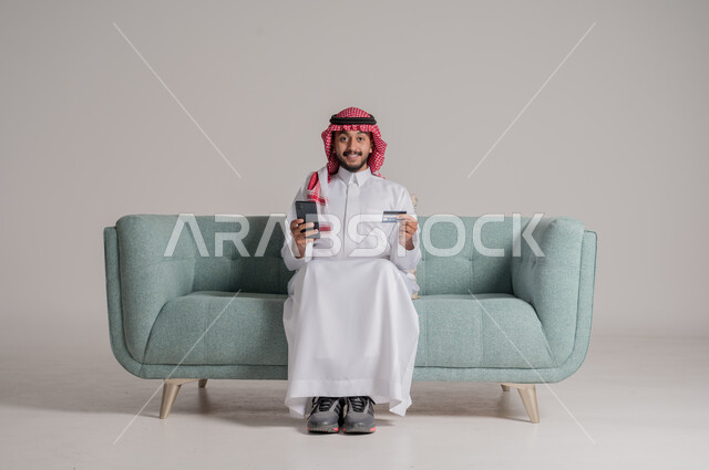 Ability to pay bills with electronic card, looking at the camera with gestures of happiness and joy, purchasing and paying through mobile programs, portrait of a smiling Saudi Arabian Gulf man wearing traditional dress sitting on the sofa holding a mobile phone and a bank credit card, gray background