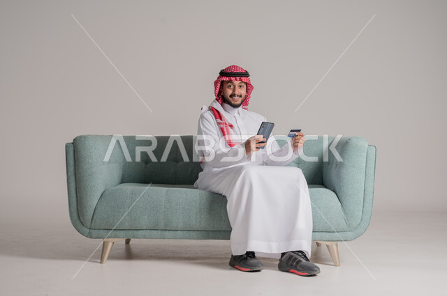 الشراء والدفع من خلال برامج الجوال، بورتريه لرجل عربي خليجي سعودي مبتسم يرتدي الزي التقليدي يجلس على الكنبة ويمسك بيده الهاتف المحمول وبطاقة الإئتمان البنكية، إمكانية تسديد الفواتير بالبطاقة الإلكترونية، النظر إلى الكاميرا بإيماءات السعادة والفرح، خلفية رمادية