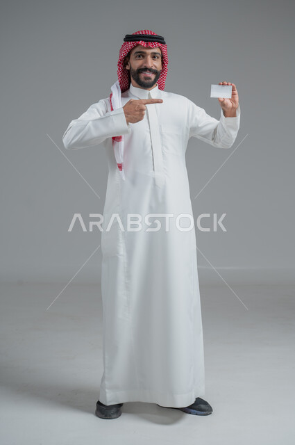إيماءات الاقناع والإعجاب بطرق الشراء الحديثة, استخدام بطاقات الائتمان البنكية, بورتريه لرجل عربي خليجي سعودي يرتدي الثوب والشماغ التقليدي يمسك بطاقة بيضاء فارغة, صورة الجسم بالكامل, خلفية رمادية