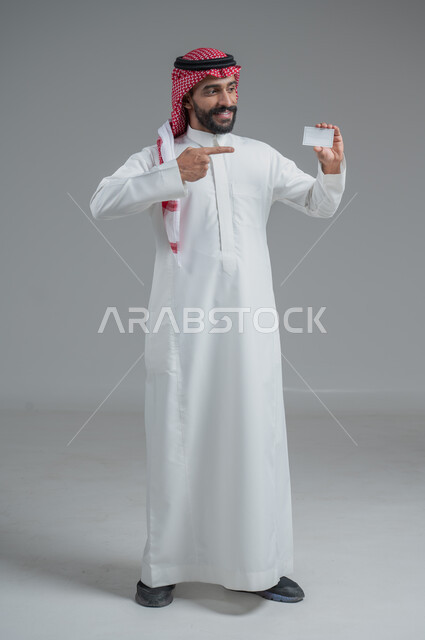 إيماءات الاقناع والإعجاب بطرق الشراء الحديثة, استخدام بطاقات الائتمان البنكية, بورتريه لرجل عربي خليجي سعودي يرتدي الثوب والشماغ التقليدي يمسك بطاقة بيضاء فارغة, صورة الجسم بالكامل, خلفية رمادية