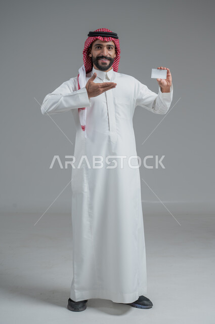 إيماءات الاقناع والإعجاب بطرق الشراء الحديثة, استخدام بطاقات الائتمان البنكية, بورتريه لرجل عربي خليجي سعودي يرتدي الثوب والشماغ التقليدي يمسك بطاقة بيضاء فارغة, صورة الجسم بالكامل, خلفية رمادية