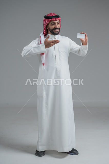 Gestures of persuasion and admiration for modern purchasing methods, use of bank credit cards, portrait of a Saudi Arabian Gulf man wearing a traditional thobe and shemagh holding a blank white card, full body image, gray background