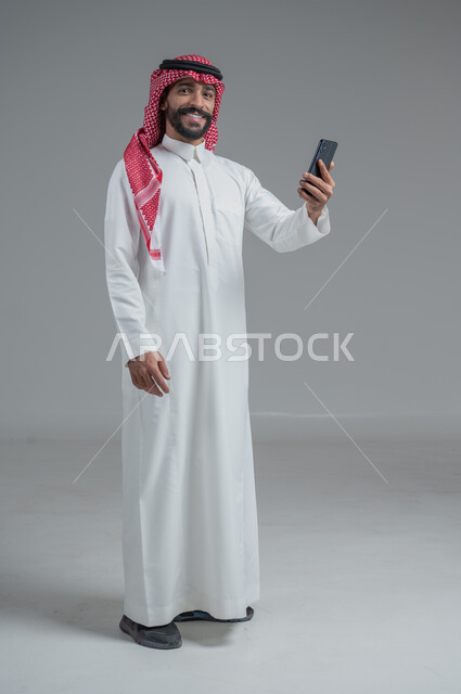 الشراء والدفع من خلال برامج وتطبيقات الجوال، بورتريه لرجل عربي خليجي سعودي يرتدي الثوب التقليدي والشماغ  ويمسك بيده الهاتف المحمول وبطاقة الإئتمان البنكية، إمكانية تسديد الفواتير بالبطاقة الإلكترونية، الجسم بالطول الكامل، خلفية رمادية