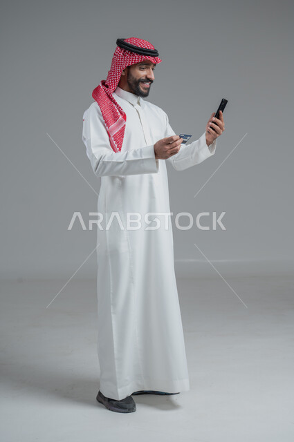 الشراء والدفع من خلال برامج وتطبيقات الجوال، بورتريه لرجل عربي خليجي سعودي يرتدي الثوب التقليدي والشماغ  ويمسك بيده الهاتف المحمول وبطاقة الإئتمان البنكية، إمكانية تسديد الفواتير بالبطاقة الإلكترونية، الجسم بالطول الكامل، خلفية رمادية