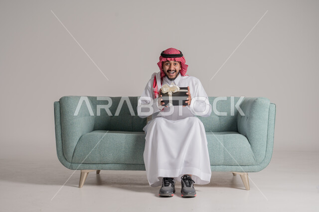 زيادة روابط المحبة بالإهداء والتقدير, التهنئات والمباركات بالنجاح والأفراح, النظر إلى الكاميرا بإيماءات السعادة والسرور، بورتريه لرجل عربي خليجي سعودي مبتسم يرتدي الثوب التقليدي والشماغ يجلس على كنبة يحمل بيده علبة هدية، أعياد ومناسبات سعيدة، خلفية رمادية