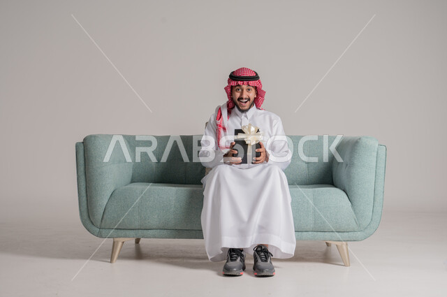 أعياد ومناسبات سعيدة، زيادة روابط المحبة بالإهداء والتقدير, التهنئات والمباركات بالنجاح والأفراح, النظر إلى الكاميرا بإيماءات السعادة والسرور، بورتريه لرجل عربي خليجي سعودي مبتسم يرتدي الثوب التقليدي والشماغ يجلس على كنبة يحمل بيده علبة هدية، خلفية رمادية