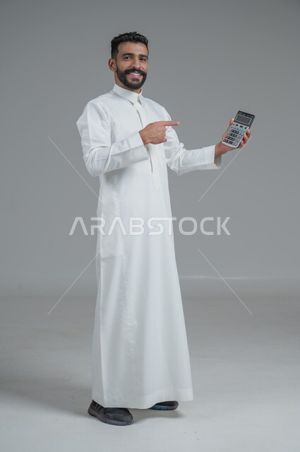 مفهوم تسهيل الاعمال والقدرة على إنجاز المهام، استخدام التقنيات الحديثة والأجهزة المتطورة ، بورتريه لشاب عربي خليجي سعودي يرتدي الثوب التقليدي يمسك بيده آلة حاسبة لإجراء عمليات الحساب، الجسم بالطول الكامل، خلفية رمادية