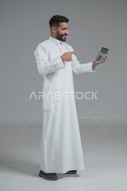 مفهوم تسهيل الاعمال والقدرة على إنجاز المهام، استخدام التقنيات الحديثة والأجهزة المتطورة ، بورتريه لشاب عربي خليجي سعودي يرتدي الثوب التقليدي يمسك بيده آلة حاسبة لإجراء عمليات الحساب، الجسم بالطول الكامل، خلفية رمادية