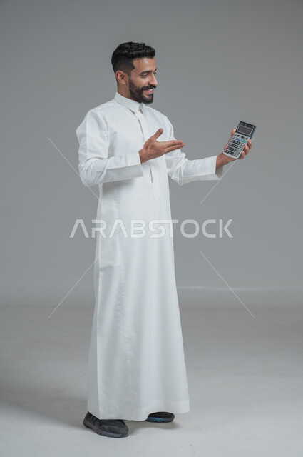 مفهوم تسهيل الاعمال والقدرة على إنجاز المهام، استخدام التقنيات الحديثة والأجهزة المتطورة ، بورتريه لشاب عربي خليجي سعودي يرتدي الثوب التقليدي يمسك بيده آلة حاسبة لإجراء عمليات الحساب، الجسم بالطول الكامل، خلفية رمادية