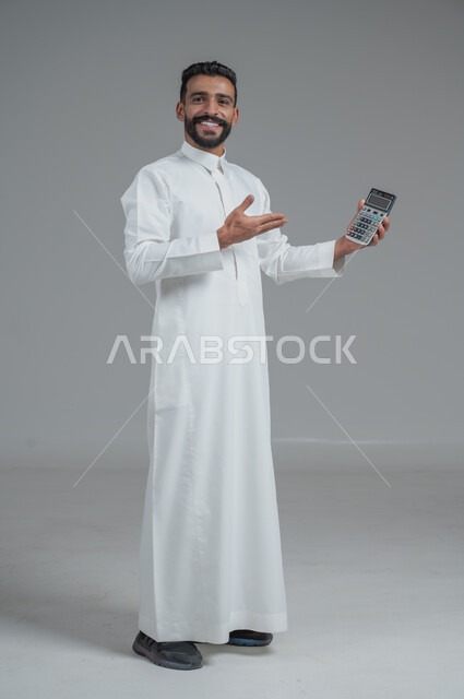 مفهوم تسهيل الاعمال والقدرة على إنجاز المهام، استخدام التقنيات الحديثة والأجهزة المتطورة ، بورتريه لشاب عربي خليجي سعودي يرتدي الثوب التقليدي يمسك بيده آلة حاسبة لإجراء عمليات الحساب، الجسم بالطول الكامل، خلفية رمادية