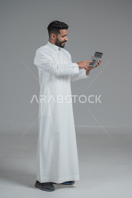 مفهوم تسهيل الاعمال والقدرة على إنجاز المهام، استخدام التقنيات الحديثة والأجهزة المتطورة ، بورتريه لشاب عربي خليجي سعودي يرتدي الثوب التقليدي يمسك بيده آلة حاسبة لإجراء عمليات الحساب، الجسم بالطول الكامل، خلفية رمادية