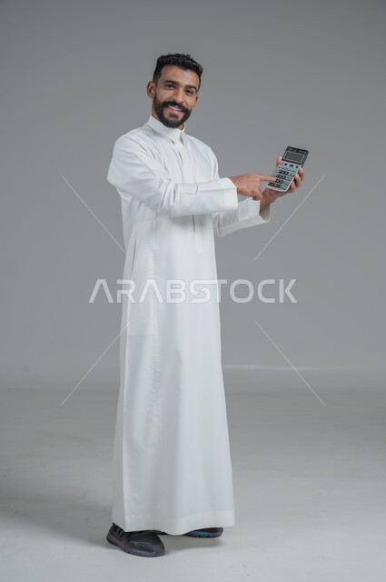 The concept of facilitating work and the ability to accomplish tasks, using modern technologies and advanced devices, portrait of a young Saudi Arabian Gulf man wearing traditional clothing holding a calculator in his hand to perform calculations, full-length body, gray background
