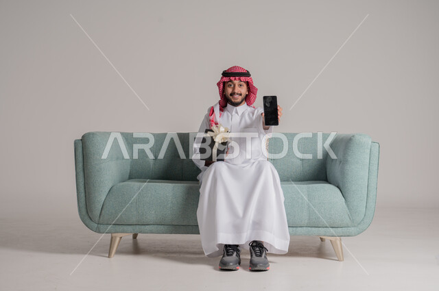 عرض شاشة سوداء فارغة، مناسبات وأعياد سعيدة، بورتريه لرجل عربي خليجي سعودي يرتدي الثوب التقليدي والشماغ يجلس على كنبة مريحة يحمل بيده صندوق هدايا والهاتف المحمول، النظر إلى الكاميرا بإيماءات السعادة والسرور، زيادة روابط المحبة بالإهداء والتقدير, خلفية رمادية