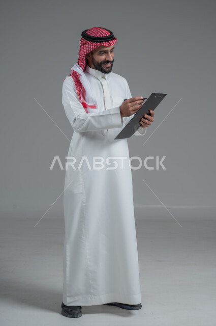 Expressions of thinking for decision making, taking notes, portrait of a Saudi Arabian Gulf man wearing traditional thobe and shemagh, holding a notebook and pen in his hands writing down information, full body, gray background