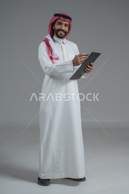 تعابير التفكير لاتخاذ القرارات, تدوين و تسجيل الملاحظات, بورتريه  لرجل عربي خليجي سعودي يرتدي الثوب التقليدي والشماغ ويمسك بيديه مفكرة وقلم يقوم بكتابة المعلومات, الجسم بالطول الكامل، خلفيه رمادية