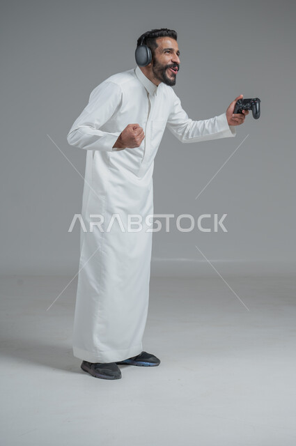 تقنيات حديثة مسلية, قضاء وقت ممتع ومسلي، التفاعل والاندماج مع ألعاب البلايستيشن، بورتريه لرجل عربي سعودي خليجي يرتدي الثوب التقليدي ويحمل بيده عصا التحكم  ويضع سماعات الرأس ويلعب بألعاب الفيديو، خلفية رمادية
