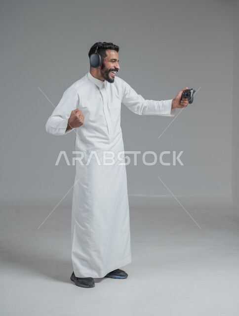 تقنيات حديثة مسلية, قضاء وقت ممتع ومسلي، التفاعل والاندماج مع ألعاب البلايستيشن، بورتريه لرجل عربي سعودي خليجي يرتدي الثوب التقليدي ويحمل بيده عصا التحكم  ويضع سماعات الرأس ويلعب بألعاب الفيديو، خلفية رمادية